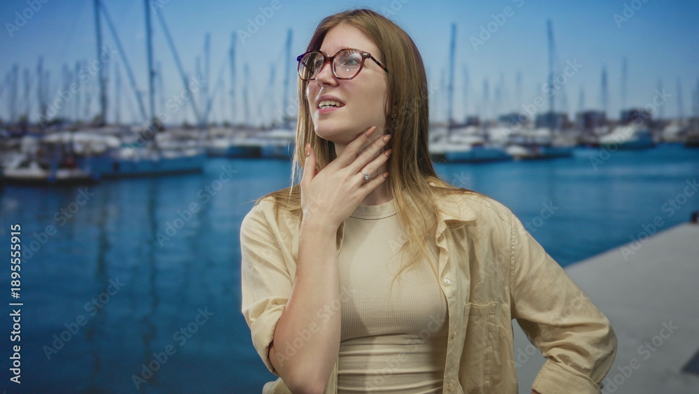 Fototapeta premium Blonde woman wearing glasses pinches nose beside boats on sunlit street pier over blue water; disgust.