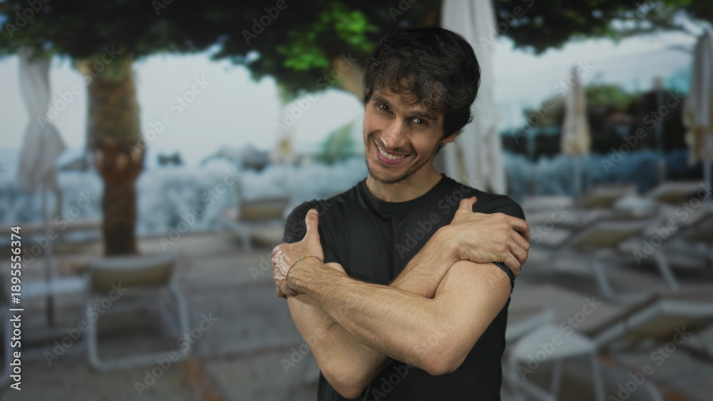 Fototapeta premium Man with bare forearms and smiling face crosses arms hugging chest at building entrance by pool chairs and umbrellas; contentment.