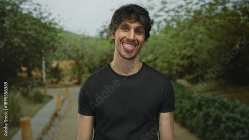 Man wearing a black t shirt sticks out tongue and tilts head with a playful smirk on a tree lined pathway in a forest setting; mischief.