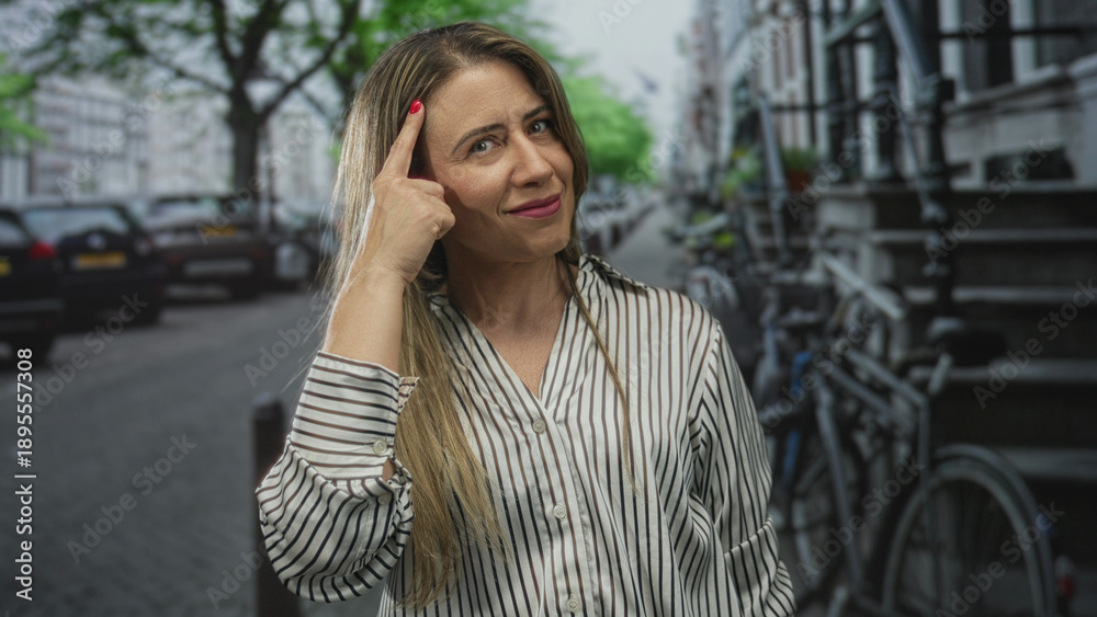 Fototapeta premium Blonde middle aged woman with long hair wearing striped shirt pointing finger to head on green street; thinking.
