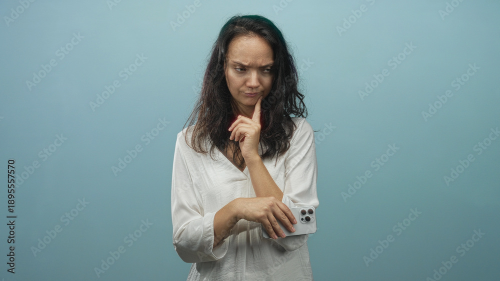Obraz premium Woman holding smartphone with finger to chin and examining phone screen in studio; uncertainty hesitation doubt.