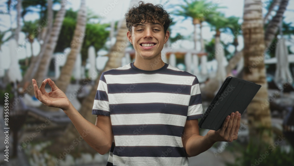 Fototapeta premium Teenage boy holding tablet and making ok sign beside palm trees and pool at resort building; relaxation mindfulness.