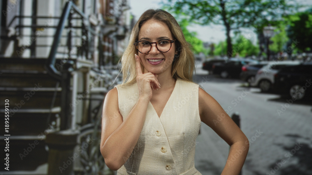 Fototapeta premium Woman with hand on cheek on street wearing glasses and sleeveless beige vest, smiling in urban outdoors; thoughtful curiosity.