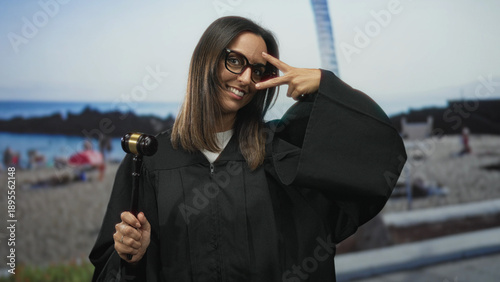 Photography Woman judge in black robe holds gavel and makes a v peace sign with fingers to her eye in studio; playful confidence