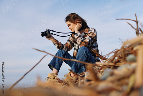Foto Photographer, woman and camera in an outdoor setting with natural light, casual plaid shirt, sitting on driftwood, capturing landscape moments with focused expression and calm mood