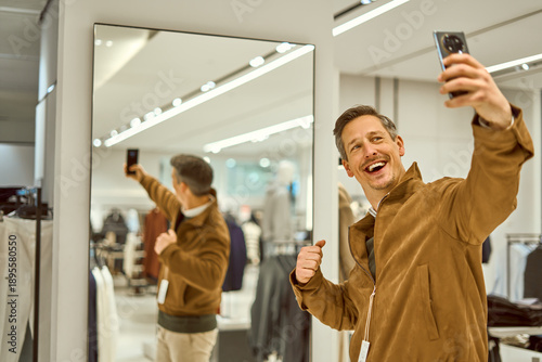 Valokuva Smiling Man Shopping in Mall Takes Selfie While Trying On Brown Jacket in Store
