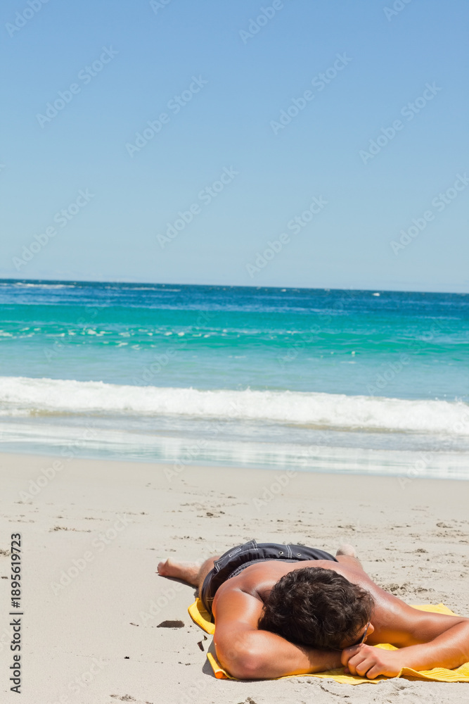 Fototapeta premium Male is lying face down on yellow beach towel on pale sand with gentle waves breaking