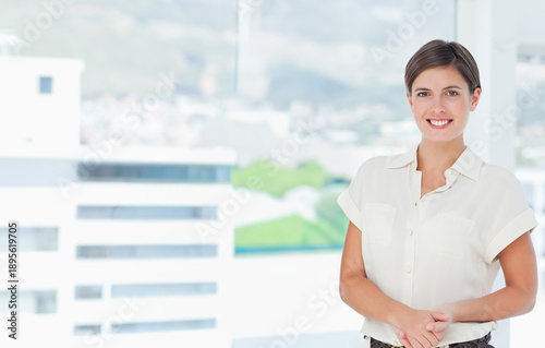 Fotografie Adult female standing by floor-to-ceiling windows at office, smiling, white blou