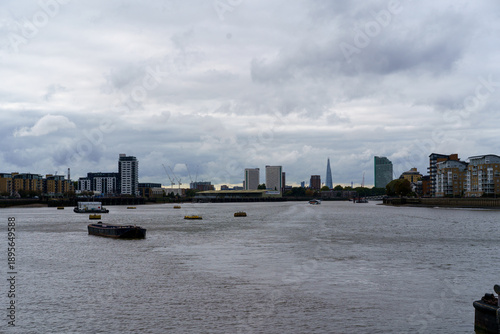 Cloudy London cityscape along the Thames, featuring the city skyline, The Shard, and boats on the river.