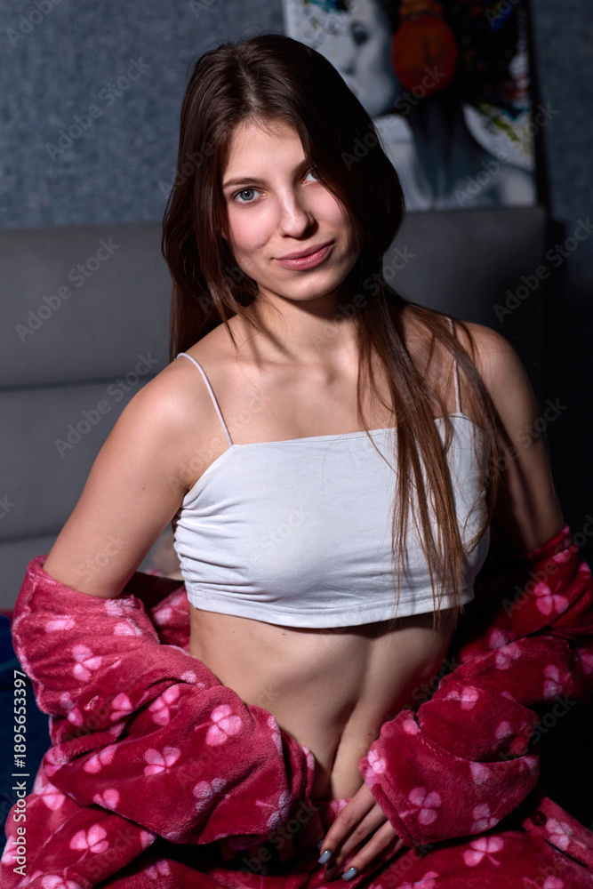 Fototapeta premium Captivating indoor portrait of a young woman, casually dressed in a top and a pink bathrobe, showcasing her playful and confident personality through a subtle yet engaging expression.