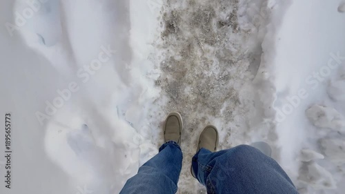 Point-of-view shot of a man walking on an un-shoveled sidewalk covered in ice and packed snow, highlighting slippery winter conditions and pedestrian safety hazards. 15033