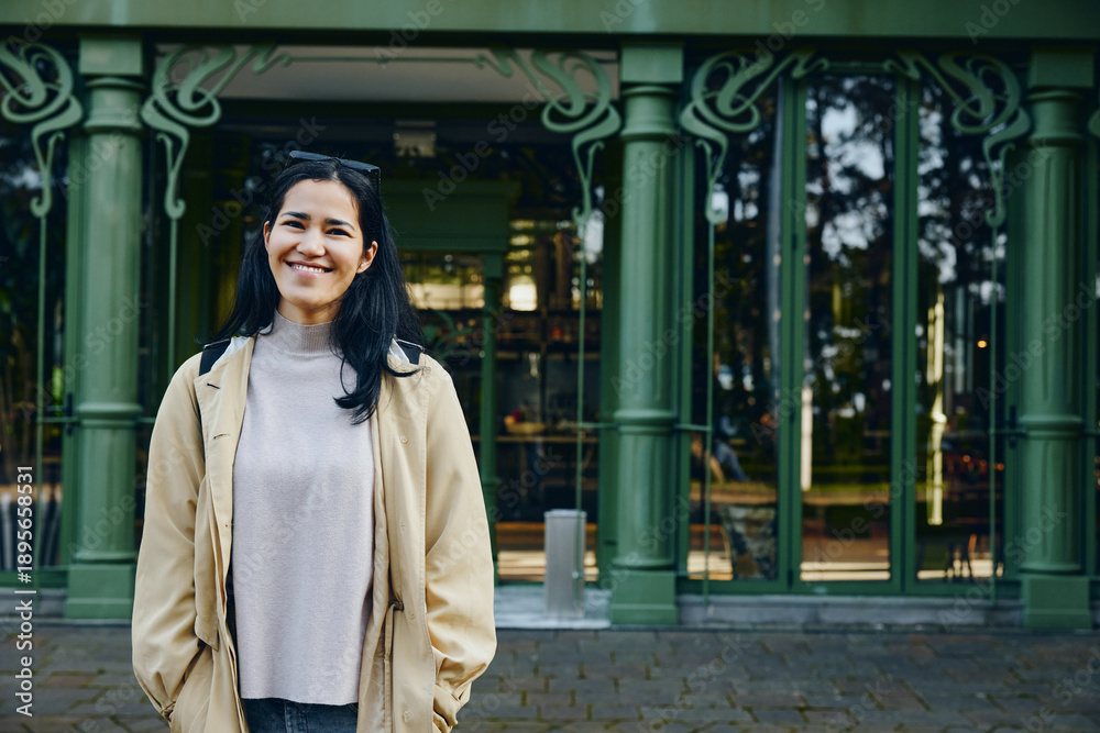 Fototapeta premium Smiling woman stands outdoors in front of a green ornate building, wearing a beige jacket and gray sweater, candid urban scene with natural light.