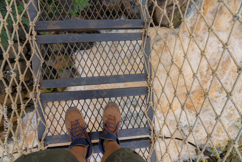 Obraz na plátně Point-of-view of hiker standing on narrow rope-and-metal footbridge suspended above churning muddy rapids