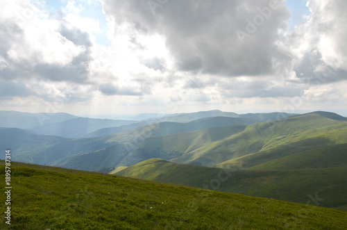 Vast green hills roll into distant mountains beneath a dramatic, cloud filled sky. Soft sunlight breaks through, creating peaceful, natural scenery. Carpathian Mountains, Ukraine 