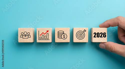 A hand carefully places a wooden block with 2026 on it next to blocks showing icons for business team growth money and success on a clean blue backgro
