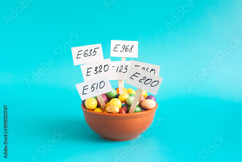 Schilderij op canvas A cup with various colored food additives in the form of granules and corresponding inscriptions and warning signs in close-up on a light background