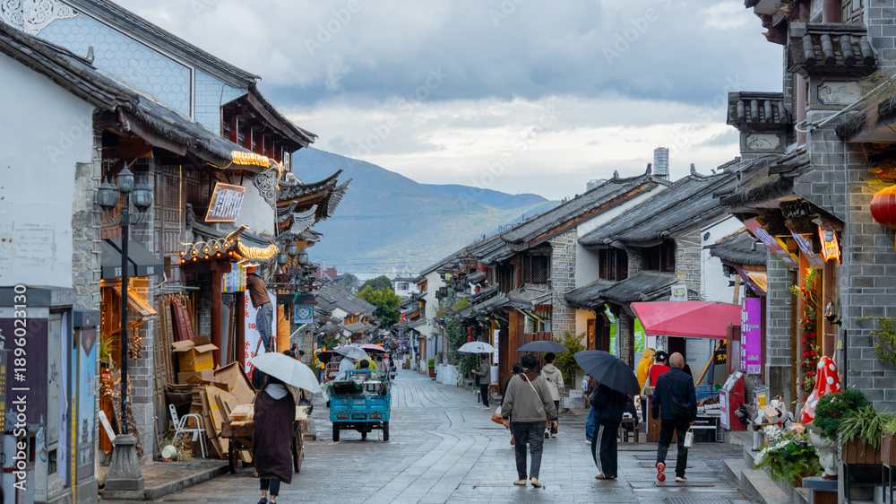 Obraz premium People with umbrellas walking through an ancient town street with traditional Chinese architecture on a rainy day.