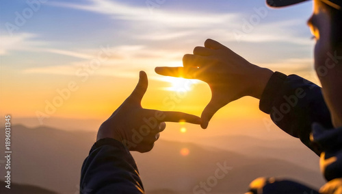 Close up shot of a man's hands making a frame gesture around the setting sun with mountains in the background during golden hour