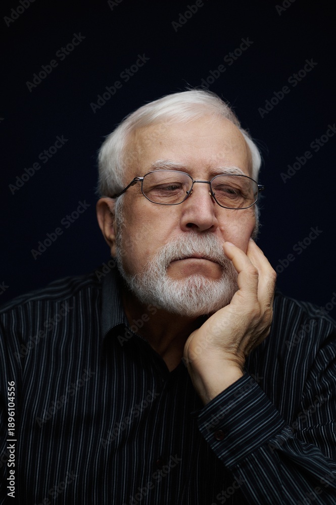 Obraz premium Older man with glasses holding his chin while deep in thought in a dark setting