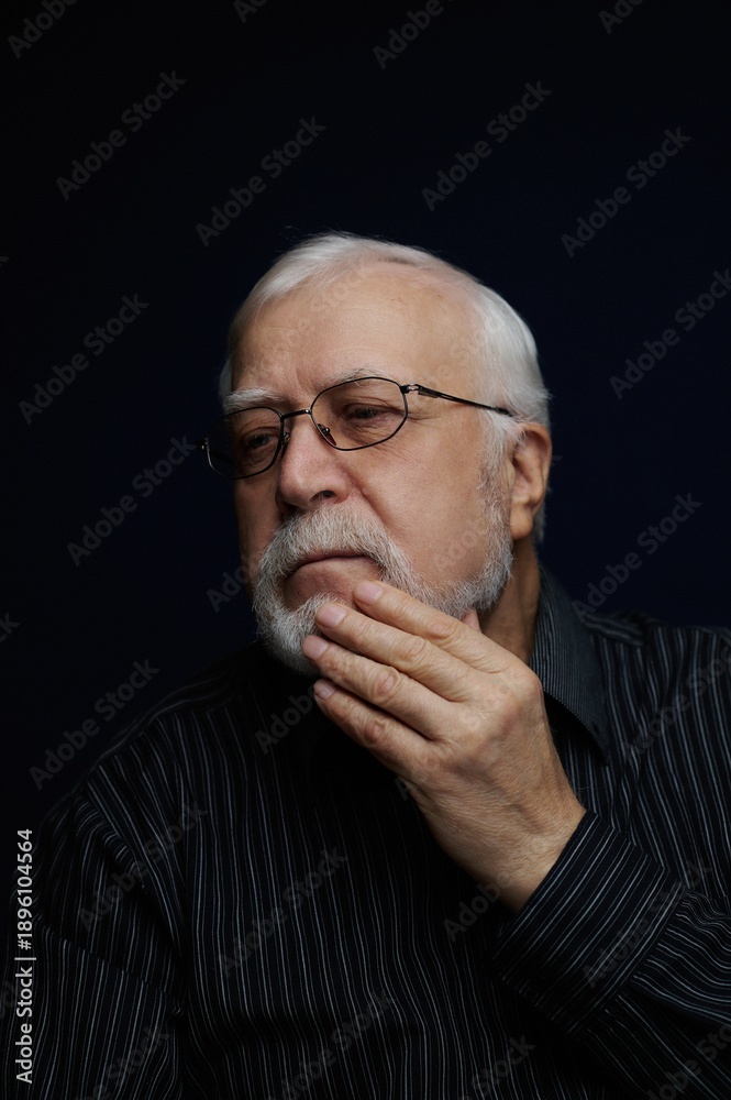 Obraz premium Older man with glasses touches his chin while thinking in a dark setting