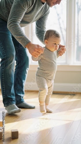 Father helping baby take first steps at home. Loving dad holding toddler hands while walking on wooden floor near window. Family lifestyle and early child development concept