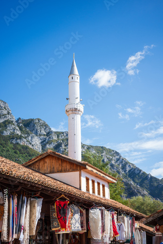 A vibrant outdoor souvenir market featuring colorful handmade textiles, rugs, and crafts displayed on a rustic wooden building, with a tall white mosque minaret rising against dramatic rocky mountains