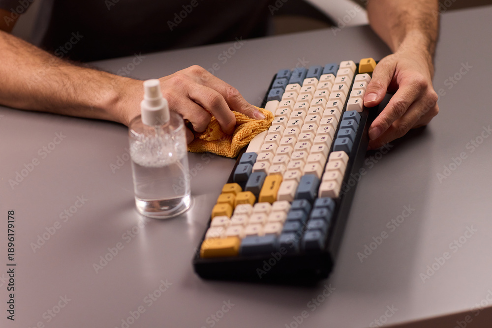 Fototapeta premium Hands cleaning mechanical keyboard with cloth, spray bottle on gray desk, closeup of keys, routine workstation hygiene, dust removal and disinfection, focused tidy atmosphere