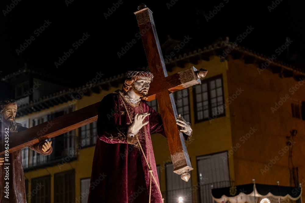 Obraz premium Jesus christ statue wearing a crown of thorns and purple robe, carrying a wooden cross, accompanied by another saint statue, during a holy week procession at night