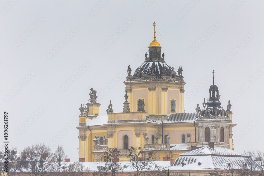 Obraz premium St George Cathedral baroque architecture view during heavy winter snowstorm. Beautiful landmark facade with golden dome under falling snow in Lviv old town. Cold cloudy evening scenic landscape.