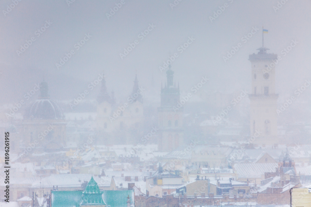 Obraz premium Panoramic view of Lviv old town landmarks under heavy snowstorm. City Hall tower, Korniakt tower and Dominican Church dome visible through blizzard. Winter urban landscape of historical center.