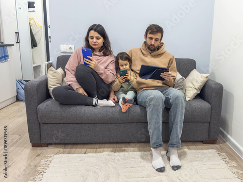 Fotografie Family sitting together on sofa while each member uses smartphone or tablet, sho
