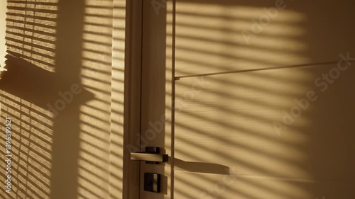Abstract pattern of light and shadow plays across a smooth wall next to a white door, illuminated by warm, late afternoon sun filtering through window blinds.