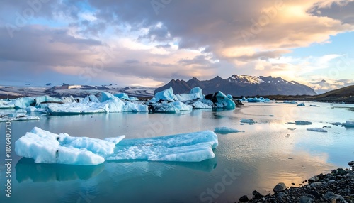 Glacial lagoon with icebergs, reflecting light. Distant mountains under a cloudy sky create a serene landscape