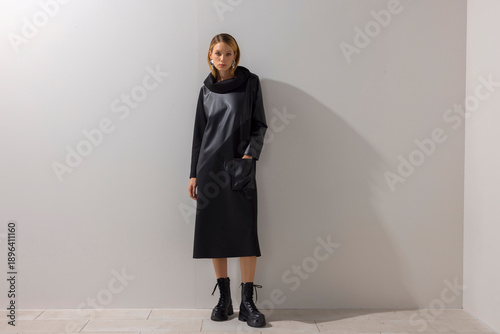 Canvastavla Full body fashion portrait of a young woman wearing a modern black dress with boots, standing against a clean studio wall