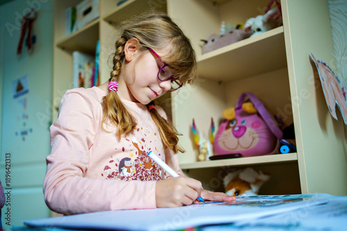 Schilderij op canvas Young girl with glasses sitting at a desk at home, drawing and learning in a cozy children room, natural light, real everyday family life and education scene