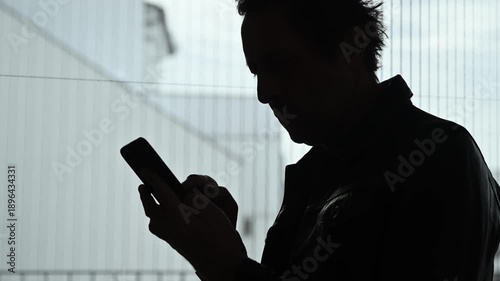 Dark silhouette of man using mobile phone next to window, symbolizing technology, isolation, modern communication, or secrecy. 4K.