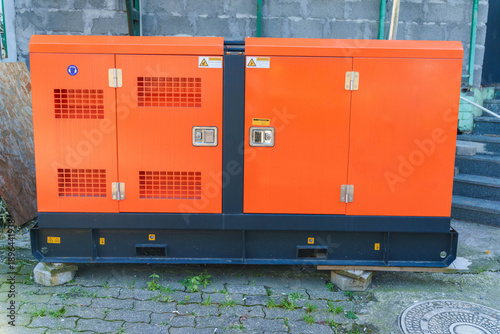 Fotografie Outdoor industrial generator with orange casing on concrete blocks near brick wa
