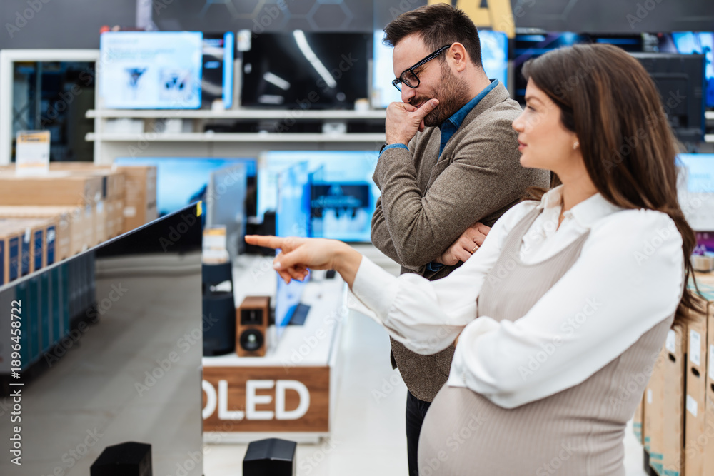 Fototapeta premium A smiling young couple shops for home entertainment, looking at a large flat screen television in a modern tech retail store department.