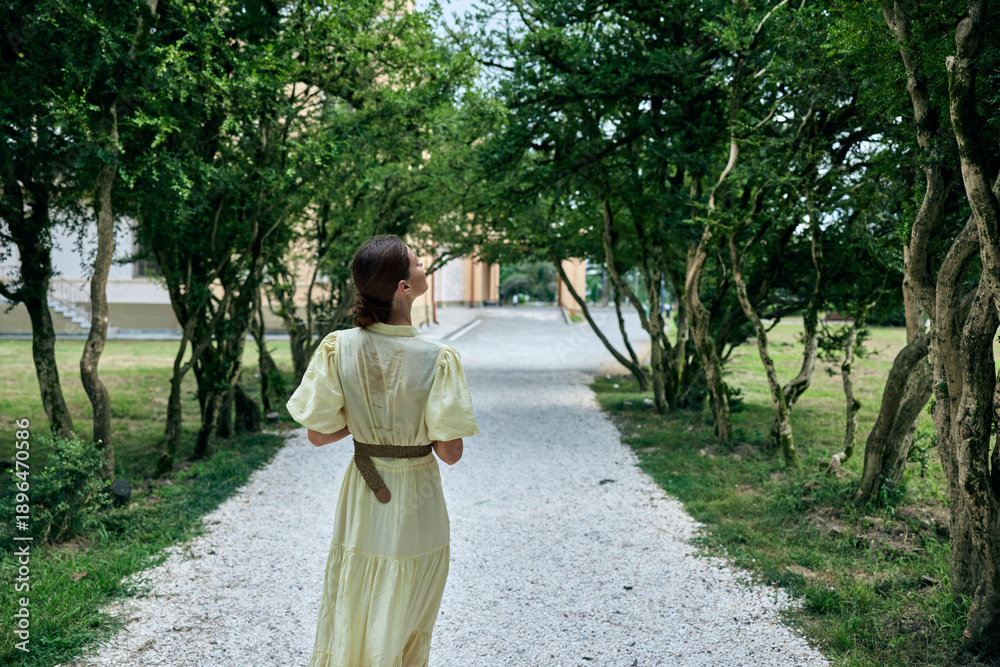 Obraz premium Woman in a pale dress walks along a gravel path under lush trees in a peaceful park, conveying calm, exploration and a timeless outdoor moment