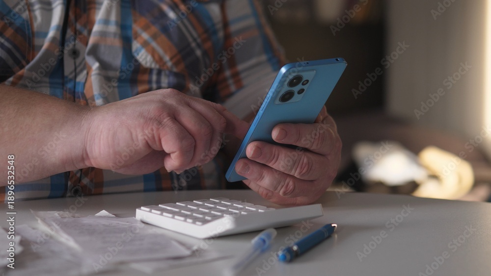 Fototapeta premium Individual looks at smartphone with one hand while using a calculator with the other. Receipts and a pen are spread out on the table in a bright indoor setting.