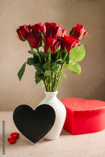Valentine's Day background. Red roses flowers in a vase, heart gift box on neutral background. Festive greeting card, flat lay
