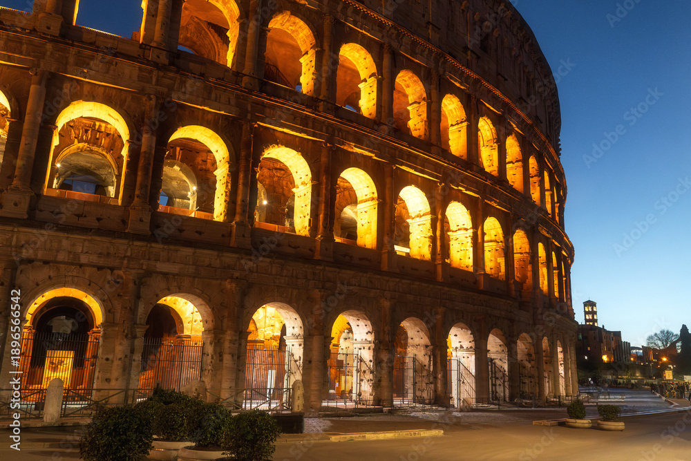 Fototapeta premium Evening cityscape with ancient walls of Colosseum in Rome, Italy. Texture of weathered travertine stone and Roman brickwork under blue sky.