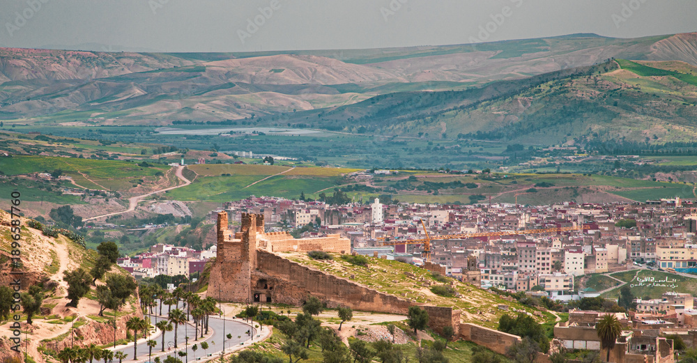 Fototapeta premium Historic walls of Fez overlooking the medina, ancient architecture and lively city, green hills in the background, contrast between heritage and natural landscape.