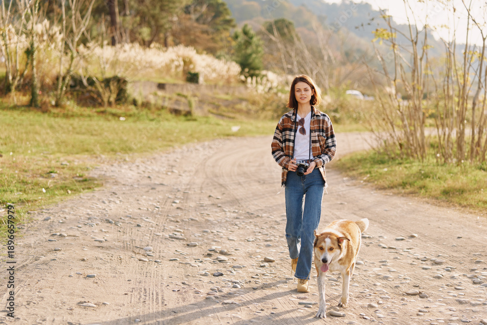 Fototapeta premium Woman walking on a dirt road with a leash dog in a rural setting, casual attire and plaid shirt, jeans, enjoying nature and daylight with a calm countryside mood.