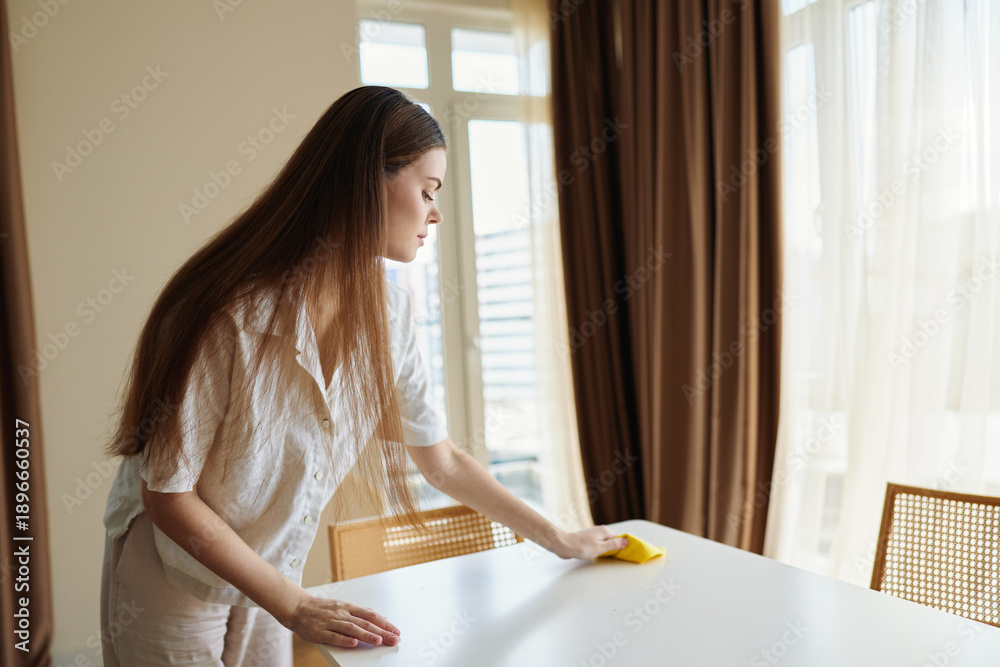 Obraz premium Woman cleaning a table with a sponge in a bright home interior, sunlight through curtains, wiping surface during morning routine for tidy, neat household and organized living space.