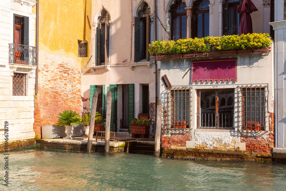 Fototapeta premium Colorful historic buildings line the edge of a canal in Venice, Italy, with arched windows, balconies, and mooring poles.