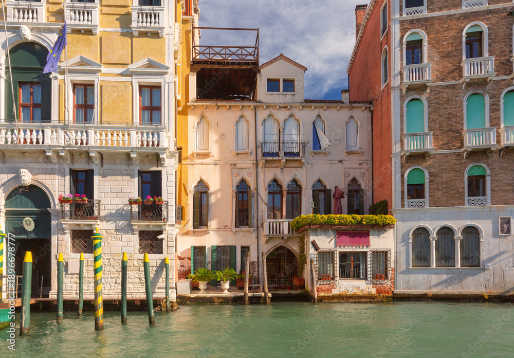 Fototapeta premium Colorful historic buildings line the edge of a canal in Venice, Italy, with arched windows, balconies, and mooring poles.