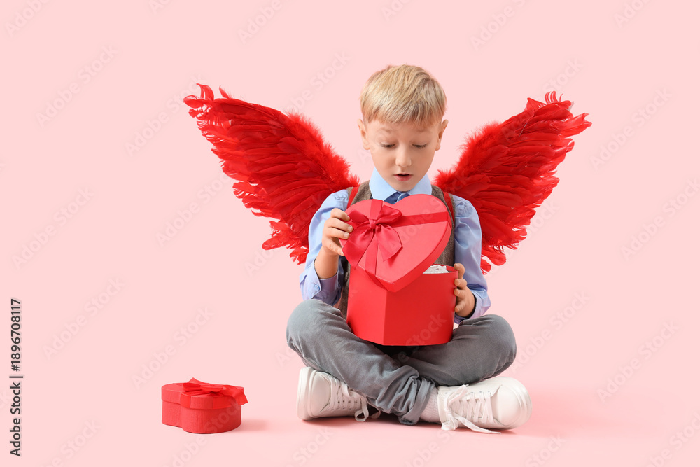 Fototapeta premium Cute little shocked boy in cupid wings with gift boxes sitting on pink background. Valentine's Day celebration