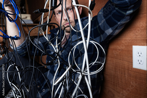 Fényképezés A stressed adult male struggles with a chaotic tangle of various electronic cables, USB cords, and chargers in a dark room