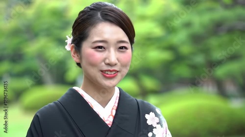 Smiling Asian woman in black kimono with floral print on shoulder against green foliage backdrop